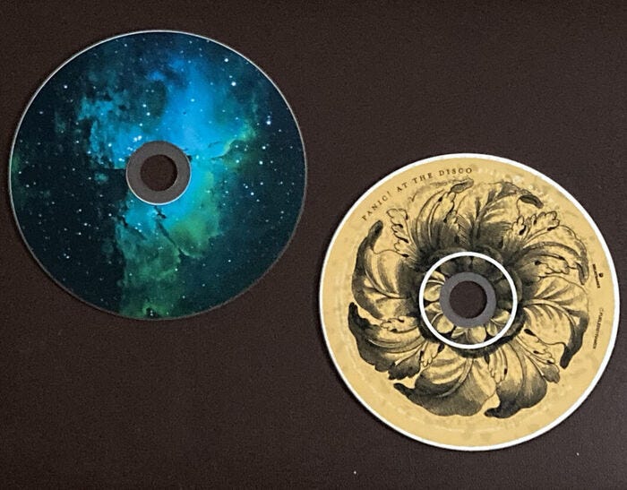 Two CDs on dark brown background; Panic! at the Disco “Vices and Virtues” on bottom right (gray outline of leaf flower on yellowed-white background and band name on portion of edge of the disc), CD at top left with blue/green nebula design