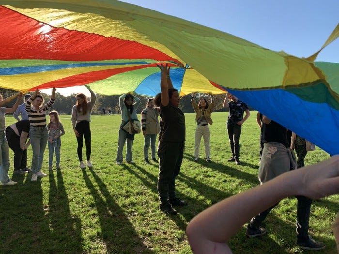 children and student volunteers playing with a rainbow parachute on a sunny day