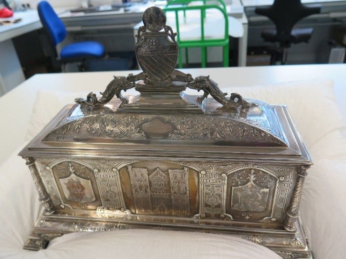 Silver casket, engraved with images of the John Rylands Library, with feet and an elaborate top piece made up of 2 sea creatures holding up a crest. The casket was presented to Mrs Enriqueta Augustina Rylands in October 1899, along with a scroll bearing the Freedom of the City of Manchester. In this image it sits in the Collection Care lab on a padded surface, waiting treatment.