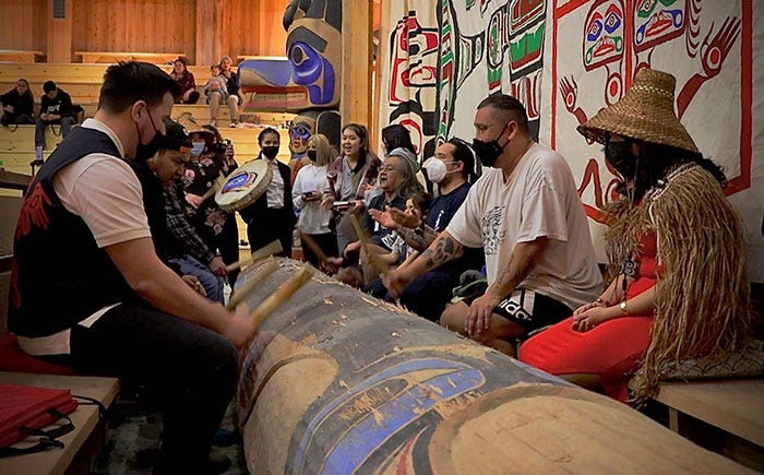 This image shows the Haíɫzaqv Nation celebrating the creation of the Haíɫzaqv Community Energy Plan (HCEP) & work toward implementation with song and dance, in a covered wooden room. All gathered around the trunk on the ground that is also painted with traditional drawings, playing with wooden sticks, singing, and wearing masks to protect themselves of the COVID-19. On the right of the photo, we can see traditional paintings with red, blue and green colours, on a white cloth in the wall.