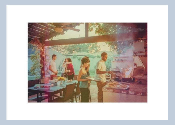 A group of young people at an outdoor BBQ and party, underneath a nice covered area with string lights and a table, and a young man is cooking.