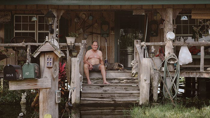 Film still from Belle River showing a grey-haired white man siting on the wooden front steps of a house wearing grey shorts and no shirt. A dog sits behind him. The house appears to be kept off the ground on stilts, and water is visible under the house, and covering the bottom steps.