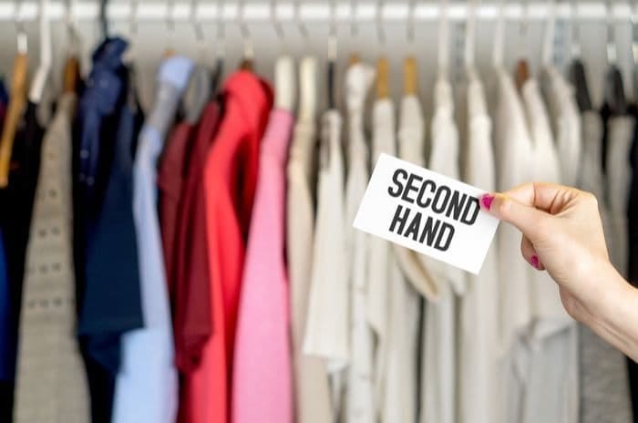 A photograph. A hand reaches from the right side of the image holding a card that says ‘SECOND HAND’. The background of the images has a blurry rack of clothes.