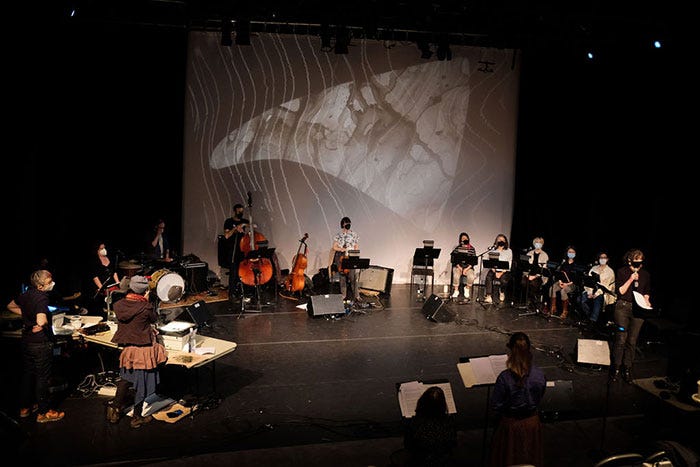 An image of a white whale fin is projected on a screen behind a black stage. Performers sit in in a circle on the stage. From left to right: two people stand at a wooden table, one person sits behind a drum kit, one behind an amplifier, one stands holding a double bass (a cello sits next to them), another stands holding a violin, and six singers sit behind black stands with sheets of music on them. One more person stands speaking into a microphone, and two more sit behind music stands.