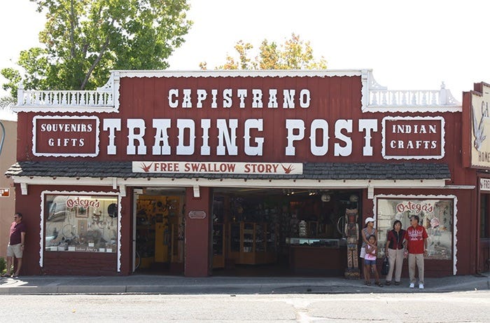 Exterior of red wooden frontier style building with white trim around the windows with a large sign in white letters that read “Capistrano Trading Post”. Two smaller signs that read “Souvenirs, gifts” and “Indian crafts” flank the larger sign on the left and right.