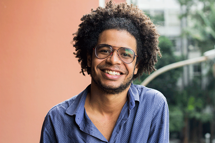 Wesley Teixeira é um homem negro, com barba e cabelos crespos e castanhos puxados para trás. Ele está sorrindo e vestindo uma camisa de botão azul. Ao fundo, há uma parede rosa e a rua, com um prédio, árvores e um poste de iluminação