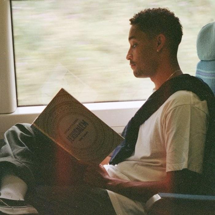Loyle Carner sitting in a train seat where the window behind him shows the countryside passing by in a blur; Loyle is holding Yotam Ottolenghi’s cookbook “Jerusalem” in his hands and reading it