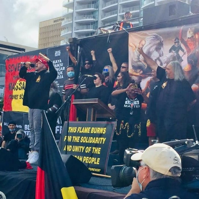 Flewnt performing live at a protest, with speakers and supporters standing behind him with their fist in the air