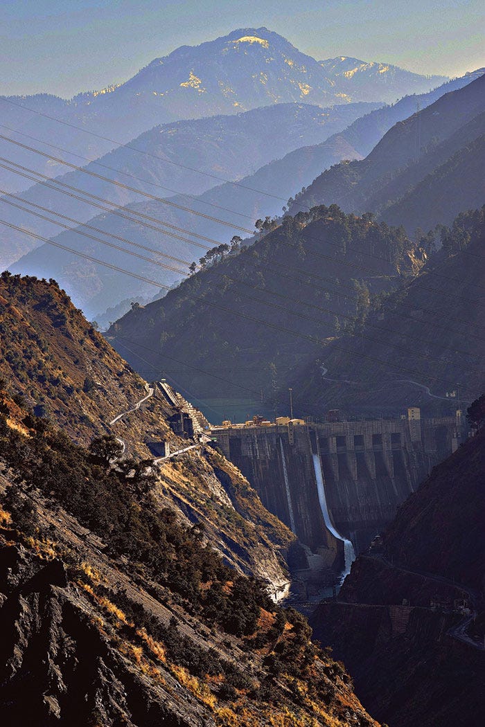 A more zoomed out view of the same photo of Baglihar Dam, showing not only the dam and the nearby hillsides, but many layers of hills in the background. The horizon is dominated by a mountain with snow on it, and many power lines are visible in the foreground.