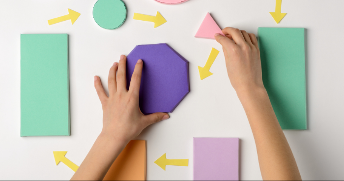 Two hands are seen arranging colorful geometric shapes on a white surface. The shapes include a purple octagon, a pink triangle, and several rectangles in green, orange, and purple. Yellow arrows, also made from paper, point in various directions, indicating a process or sequence. The arrangement suggests an exercise in design, planning, or problem-solving.Female hands precisely arranging a project plan with sticky notes and arrows.