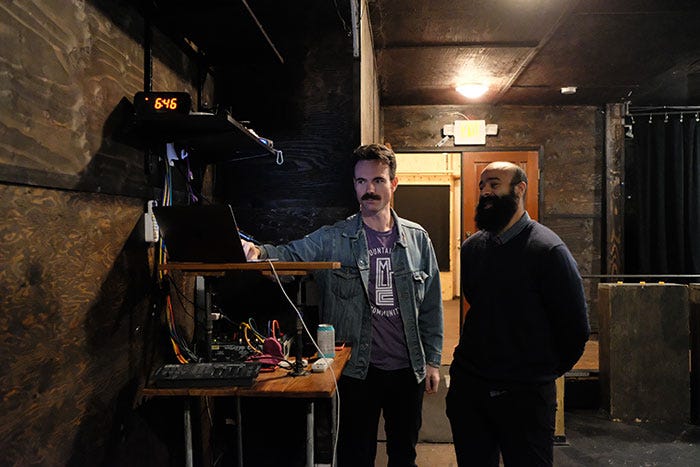 In a comedy theatre clad in plywood, two men prepare for a comedy show. One, wearing a light denim jacket, points to a computer screen, while the other, wearing a dark blue sweater, laughs.