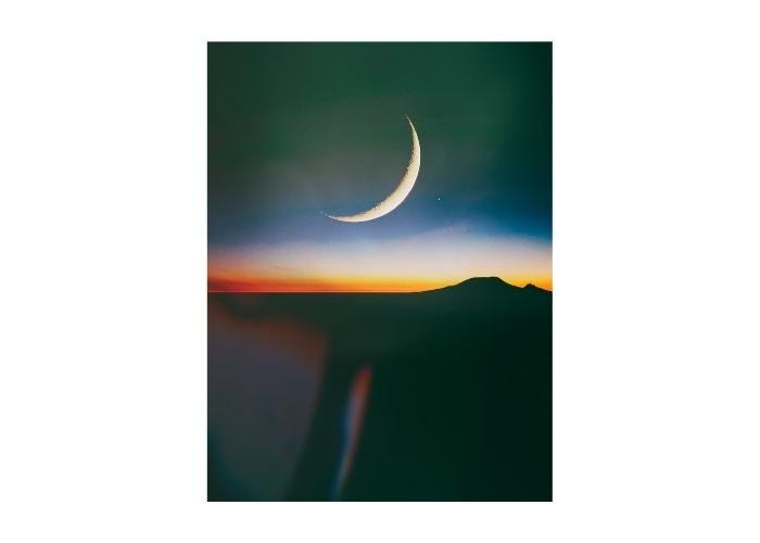 A digital photo collage showing a sliver of moon rising above a desert mountain range. The photo is almost all black except the moon and the sunset colors in the horizon