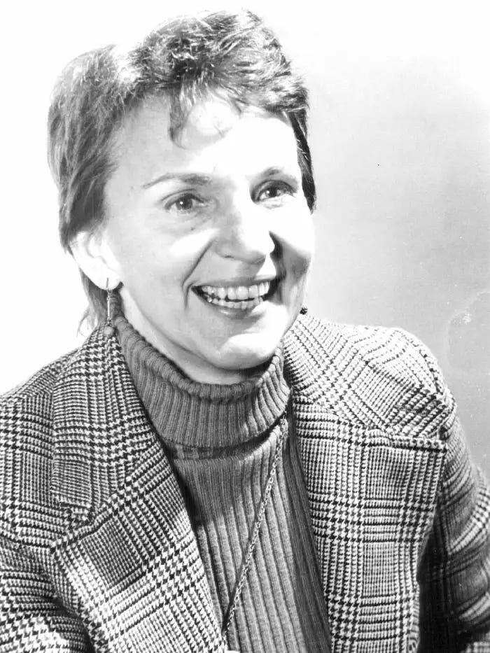 A woman with short ahir and a tweed jacket smiles brightly, looking engaged, at the camera