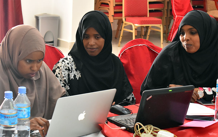 Three women sit at a table peering at two laptops placed before them.