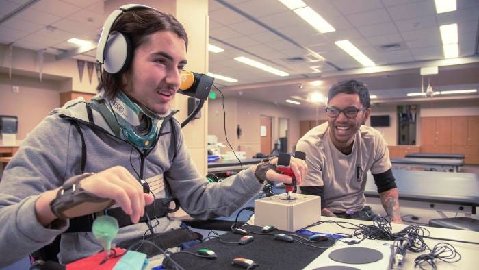 A disabled person on the left enjoying a game using the Xbox Adaptive Controller with another person on the right.