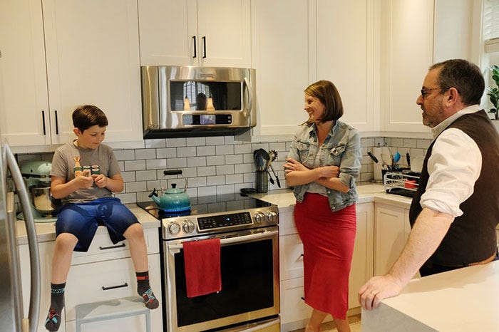 A young boy wearing a grey t-shirt and blue shorts, a woman wearing a denim jacket and a red skirt, and a man wearing a white shirt and black vest, stand in their kitchen. A blue kettle is heating on an induction stove in between them.