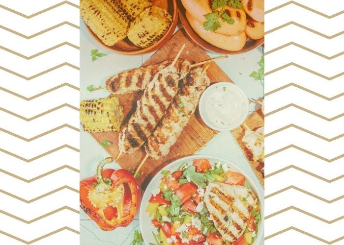 A cook-out and BBQ table spread featuring grilled and roasted corn, grilled sausages and chicken, with sauces and salad and grilled peppers.