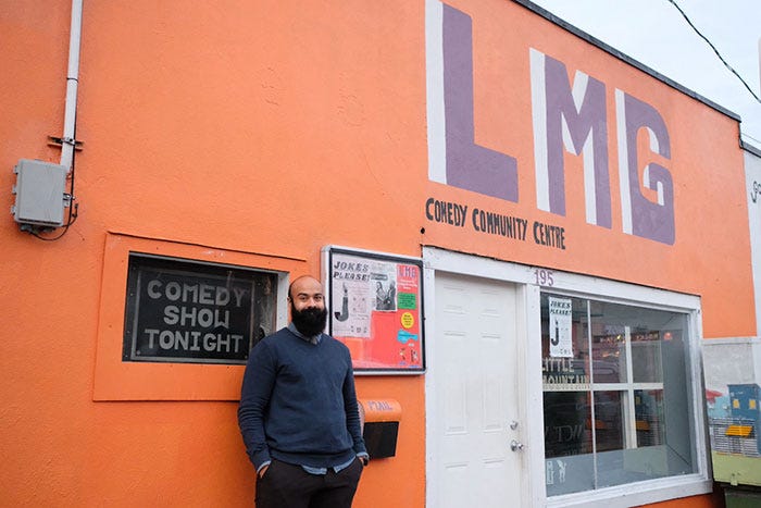 A tan-skinned man with a black beard wearing a blue sweater stands smiling outside a bright orange building with a purple “LMG” painted above its front door.