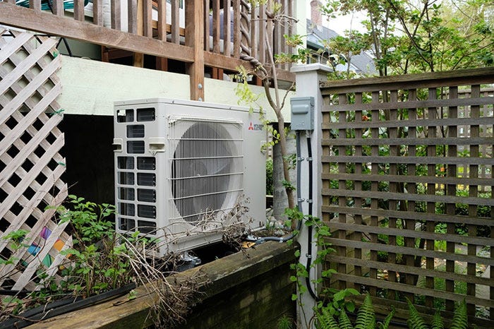 A heat pump, which is a beige-coloured rectangular machine with a circular fan, sits in a wooden-fenced side yard lush with plants.