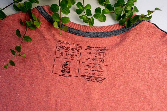 The neck-line tag of an orange Patagonia t-shirt flipped inside out, with ivy draped over the top. The tag lists sizing and material information, including recycled plastics.