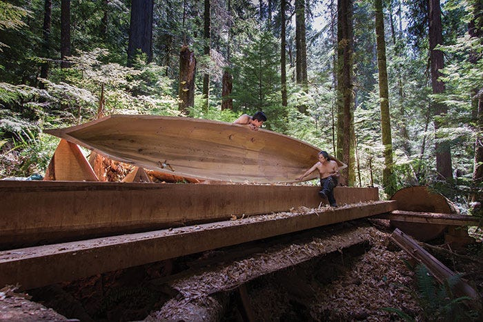 In the middle of the image, two men working with wood are shaping a traditional dugout canoe, surrounded by the green of the forest and its tall trees.