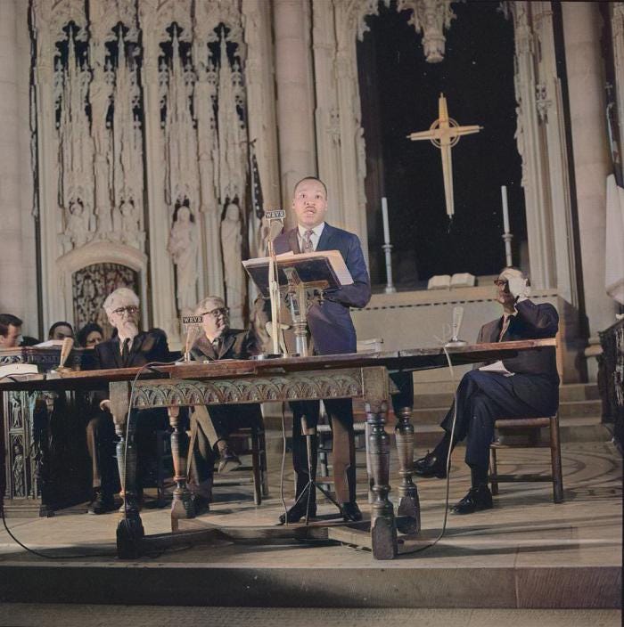 Dr. Martin Luther King stands at lectern at Riverside Church, flanked by men seated on either side
