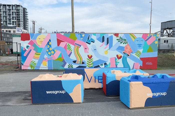 A colourful, abstract mural of shapes and faces on a wooden board in a construction site. Concrete blocks have been painted in matching colours with the logo of Wesgroup, a prominent Vancouver real estate developer.