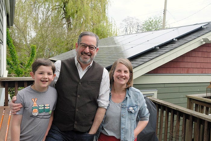 A young boy wearing a grey t-shirt, a man wearing a white shirt and black vest, and a woman wearing a denim jacket standing in front of their garage, which has solar panels on its roof.