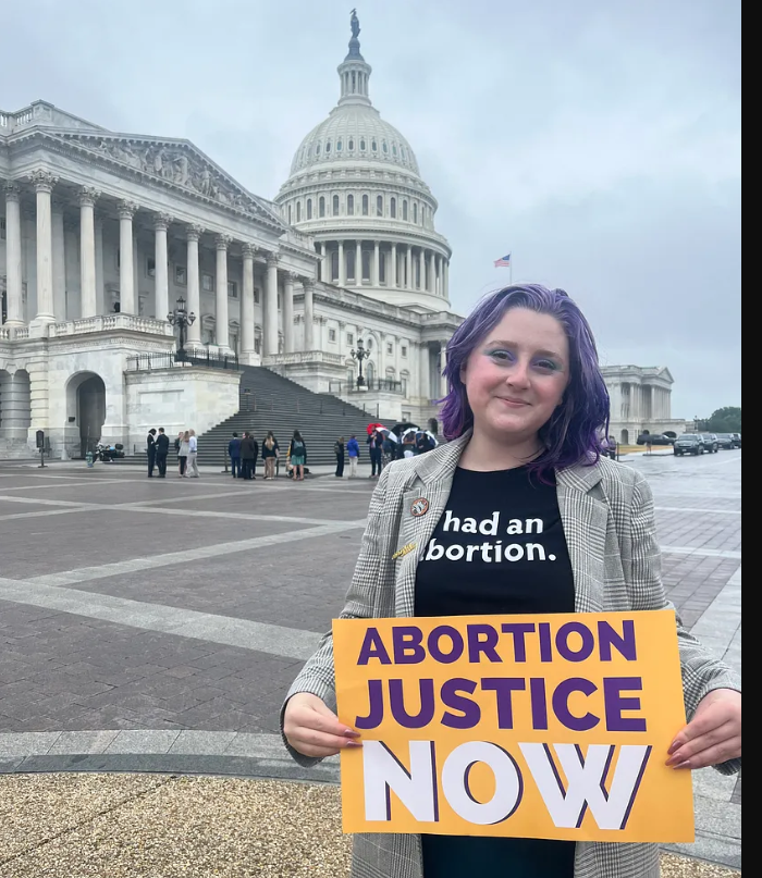 A photo of Lizzie, a White woman with purple hair. She is wearing a T-shirt that reads “had an abortion,” and holding a sign that says “ABORTION JUSTICE NOW. The Capitol building is in the background.