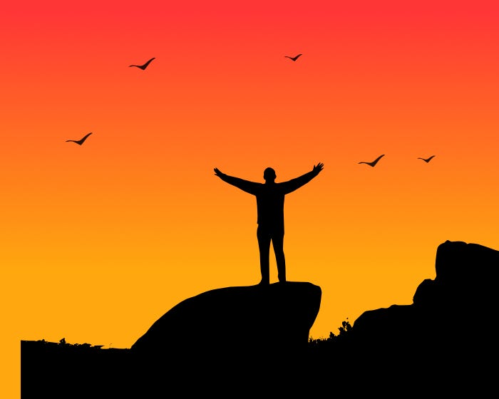 Image of a man standing at the top of a mountain at sunset, with his arms open wide, with birds flying above.