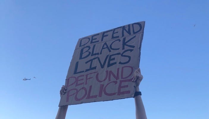 Hands thrust a protest sign reading “Defend Black Lives Defund Police” into the sky, as an LAPD helicopter flies overhead.