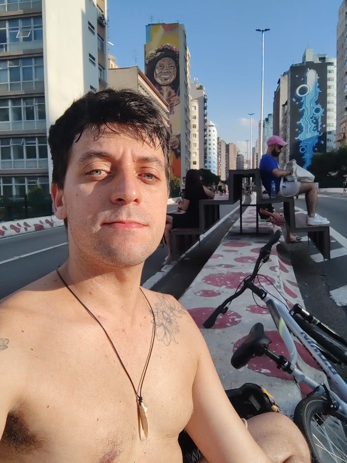 Homem branco tirando uma selfie, sem camisa, com o sol batendo na pele, usando colar de prancha de surf, sentado no meio de uma pista, com a bicicleta posta deitada de lado, com outras pessoas ao fundo sentadas em um banco.