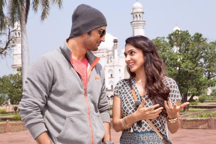 Sushant Singh Rajput is pictured in his Dhoni film attire — a grey beanie and a grey jacket. He walks beside Kiara Advani who plays Sakshi Rawat is pictured a a grey kurti. They are talking and the Taj Mahal is visible in the background.