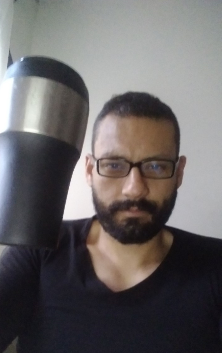 Omar is celebrating his love for coffee, by holding up a silver thermos. Omar wears glasses and a black t-shirt.