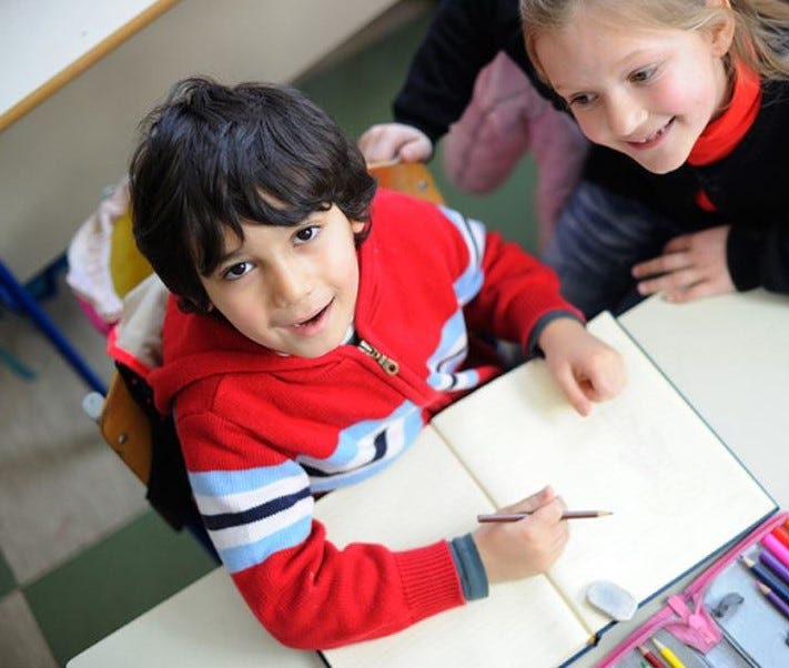 A small boy is sitting in front of a notebook with a pencil in his hand. He looks up at the camera with a smile. The girl next to him smiles at him admiringly.