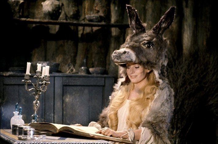 A beautiful princess sits in a humble cottage perusing a large book, but her lovely looks are hidden beneath a cloak made from a donkey’s pelt — including its head for a hood!