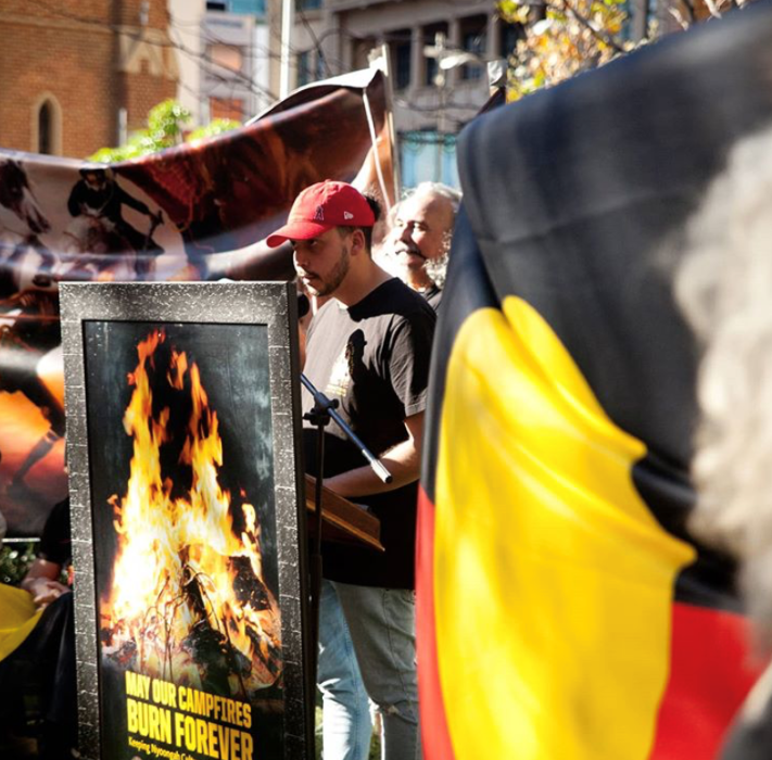 Next to the Aboriginal flag, Flewnt stands on a podium speaking. Infront is a sign stating “May our campfires burn forevever”