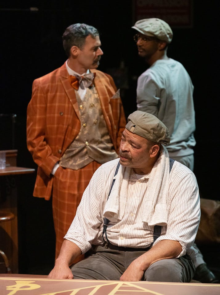 A man in an orange suit with gray hair, a mustache, and olive skin stands looking at a man in a gray sweatshirt with brown skin and glasses. Another man with brown skin and a mustache sits in front of them with a towel around his neck, wearing a hat.