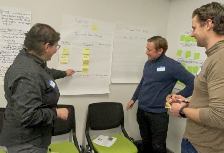 In a corner with three chairs and large white papers with handwritten text on the wall, one person stands and points to a post-it note on a large white paper with handwritten text and other post-it’s while two city employees stand and look at the post-it, listening.