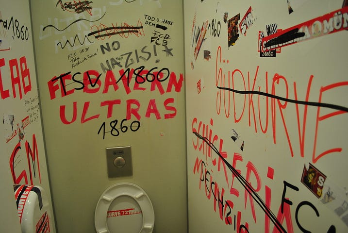 A public bathroom with boisterous comments written on the walls. Most text is not in English, but the phrase “No Nazis!” does appear.