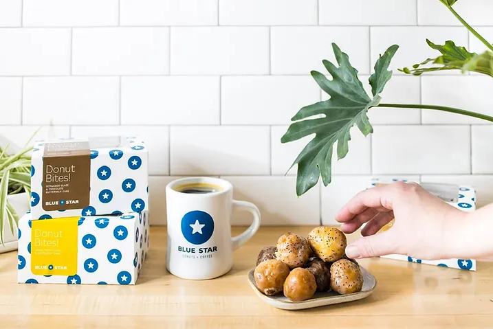 A picture of a hand reach for donut holes on a place in front of a Blue Star coffee mug.