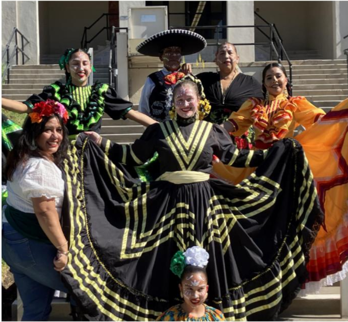A group of seven people. There are three women in a traditional flokorico dresses and their makeup is done make their faces resemble skulls. There is a man in the back dressed in charro. Finally there are two women and a little girl posing with the flokorico dancers.
