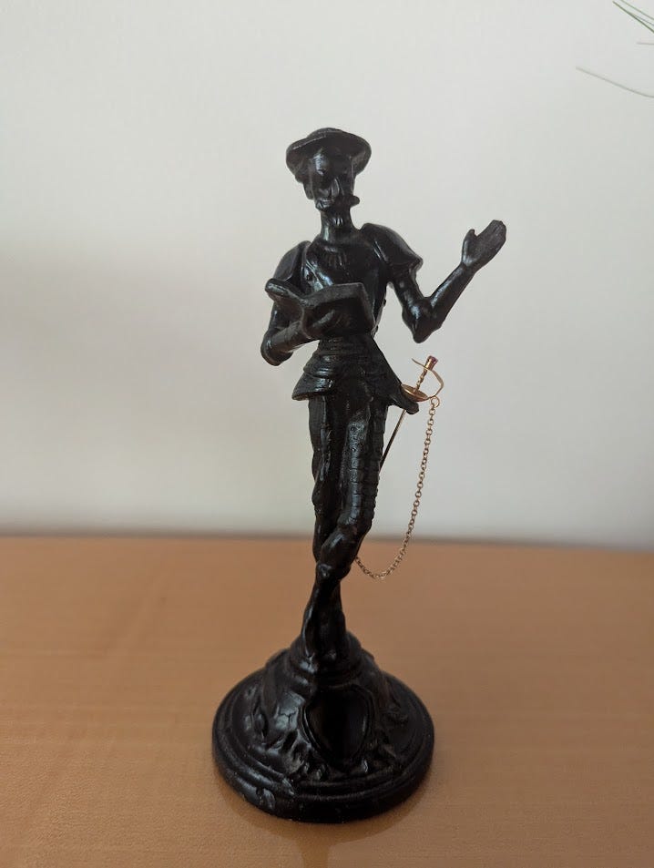 A metal figurine of Don Quixote, posing as if reading out loud from a book. He is wearing a golden, ruby pummeled sword.