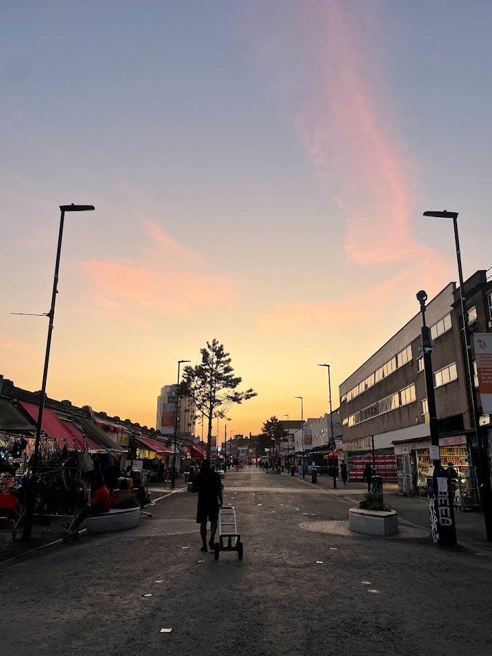 A picture of a market road in London in the evening with few people walking around, with a beautiful summer sunset in the background
