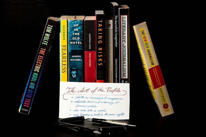 The Art of the Profile: an assortment of non-fiction books are displayed with a handwritten note and two fountain pens. The note reads: “The Art of the Profile — starts in newspapers & magazines — celebrates the lives of ordinary & famous people — can read like a novel — and become a book in its own right.”