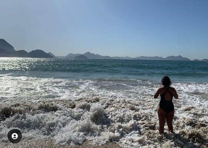 Praia de Copacabana, é possível ver morros ao fundo. É um dia ensolarado, mas o sol não está na foto. O mar está com ondas na beira, daquelas com espuma. Uma mulher de maiô e de costas pra câmera está entrando na água.