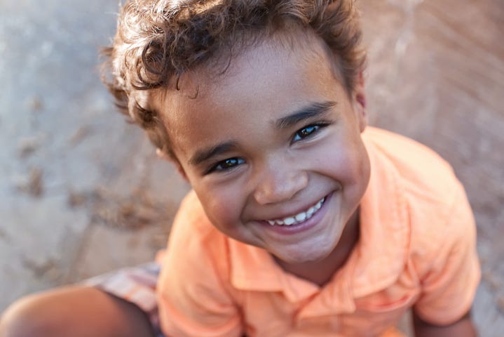 A young boy in an orange polo smiles at the camera positioned above him.