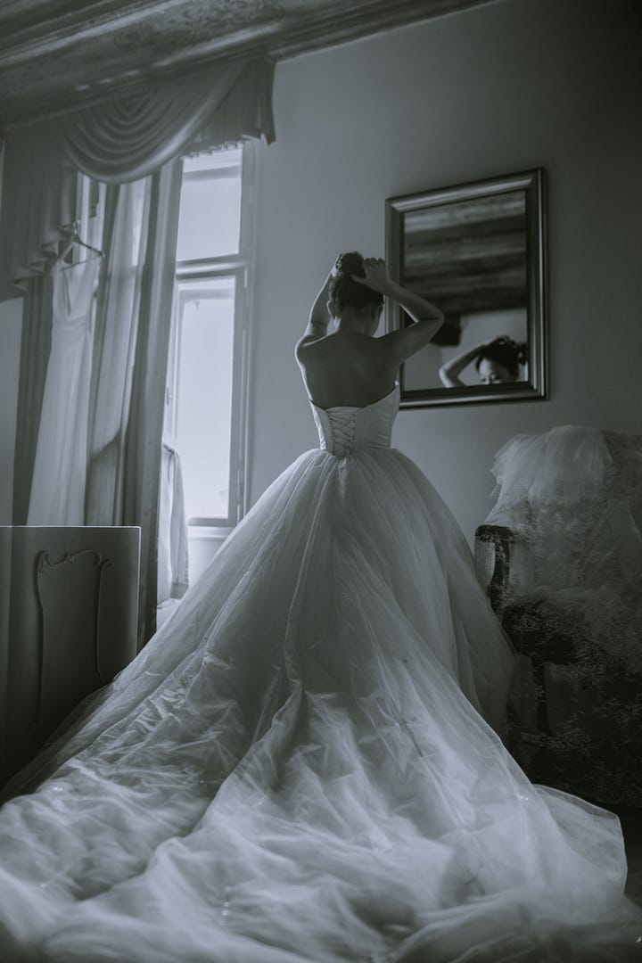 A black and white of a bride from the back, she is looking in a mirror working on her hair.