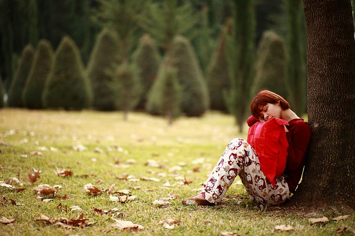 Young girl sitting against a tree, holding a red pillow in her lap