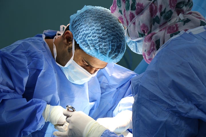 A male surgeon faces us as he holds foceps in his right hand, mask and cap on his head. We see him from the abdomen up. There is a nurse facing away from us in the foreground. Colorectal cancer incidence is rising among young people.